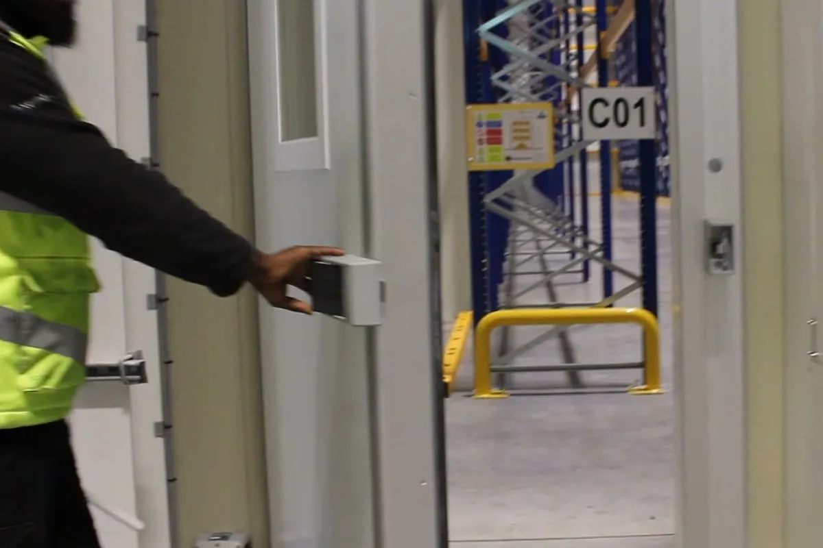 A JTF engineer fitting a door sensor to the frame of a cold store door, with racking in the aisle beyond