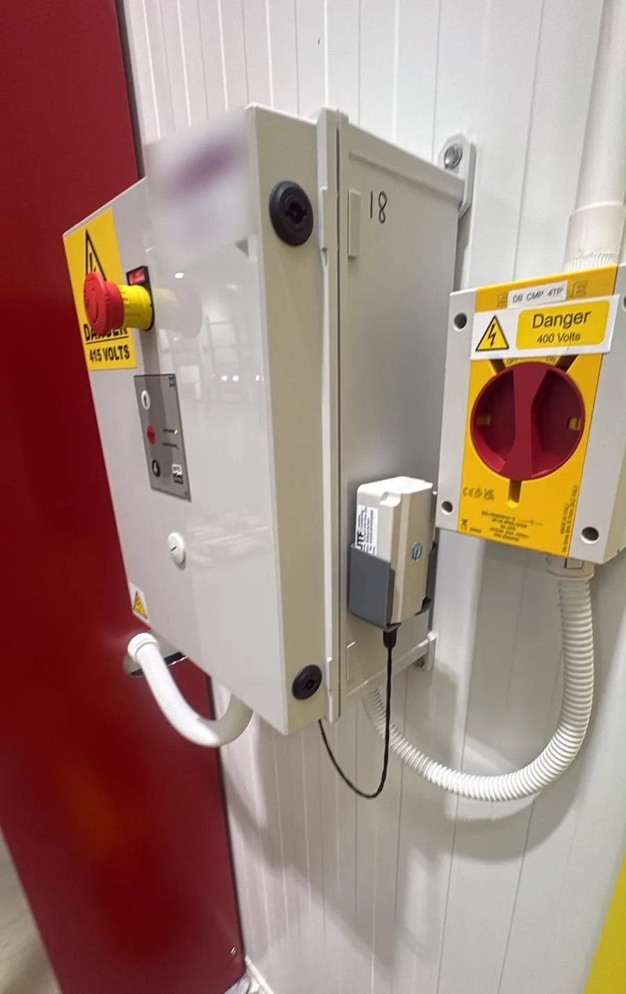 An industrial door control panel with an emergency stop and electrical warning labels, an isolator switch beside it, and a white Sematics Node+ sensor in a grey holder mounted just outside the panel, its cable running down the wall.